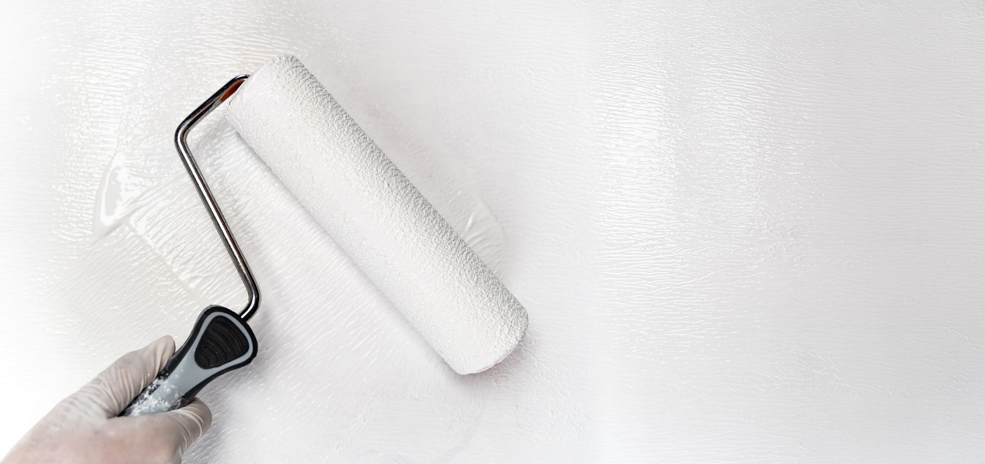 Eine Hand in einem Handschuh benutzt eine Farbrolle, um weiße Farbe auf eine weiße Oberfläche aufzutragen.