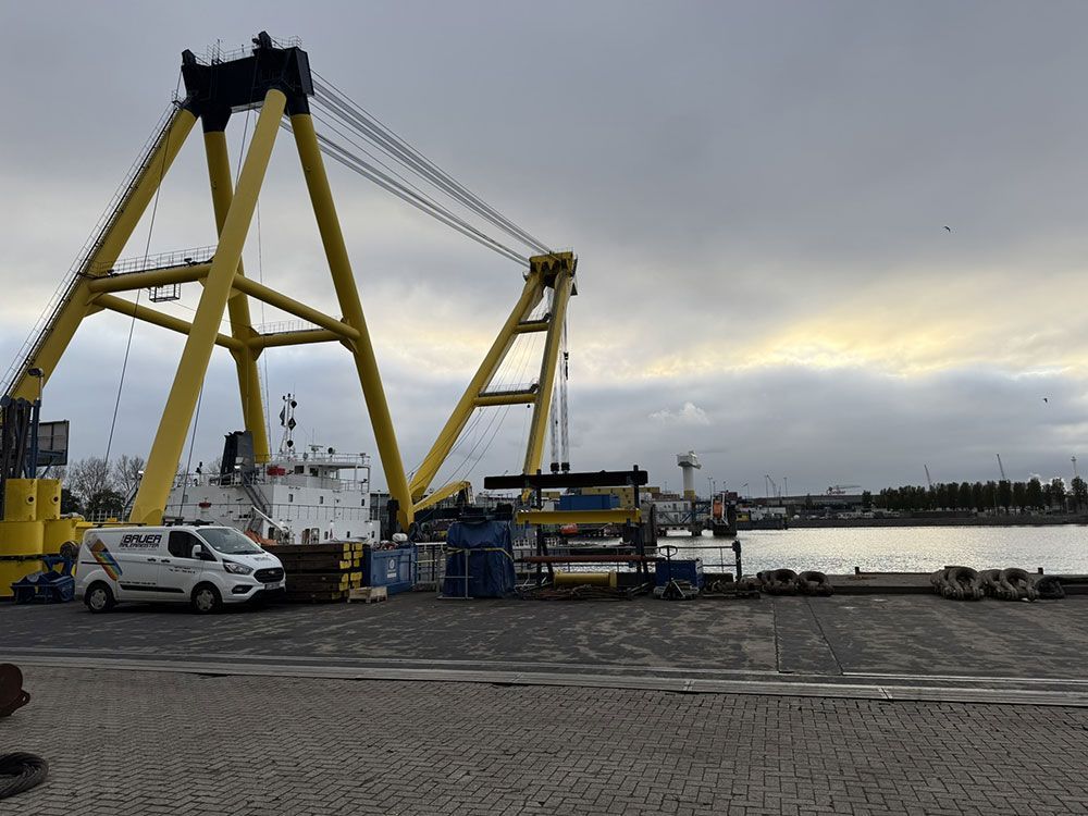 Ein großer gelber Kran und ein Schiff im Hafen, bedeckter Himmel. Ein weißer Lieferwagen steht in der Nähe.