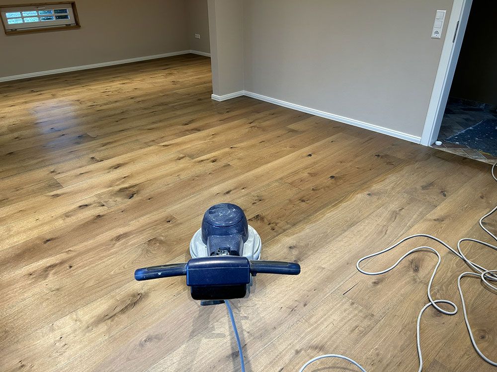 Bodenschleifmaschine auf hellbraunem Holzboden, Zimmer mit beigen Wänden und weißen Zierleisten.