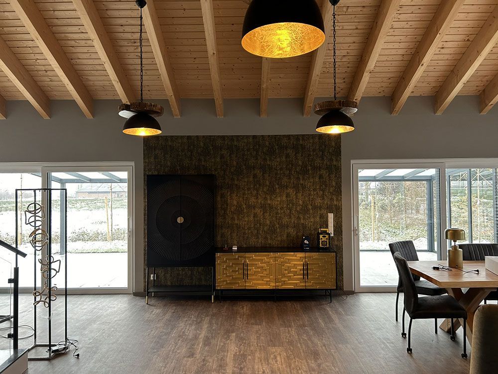 Modernes Interieur mit Holzdecke, dunklen Möbeln, goldenen Akzenten und großen Fenstern mit Blick auf ein Feld.