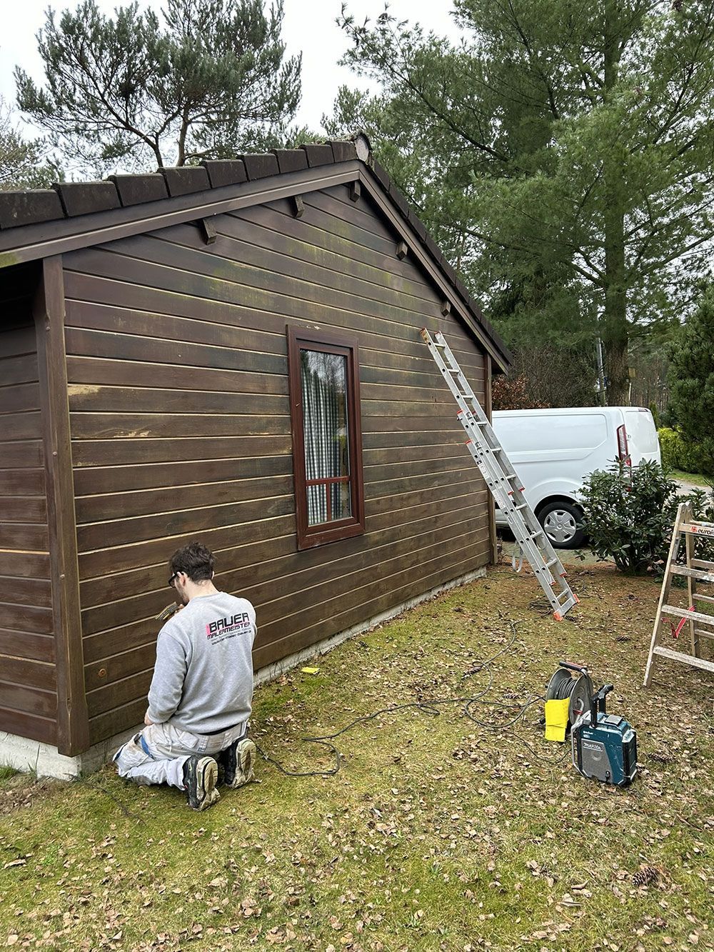 Ein Mann streicht einen braunen Holzschuppen; im Hintergrund sind eine Leiter und ein weißer Lieferwagen zu sehen.
