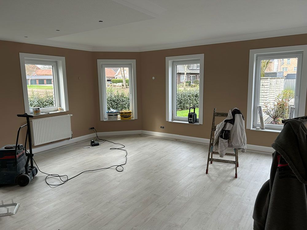 Zimmer mit beigen Wänden, vier Fenstern und hellem Holzboden. Eine Leiter und Ausrüstung sind vorhanden.