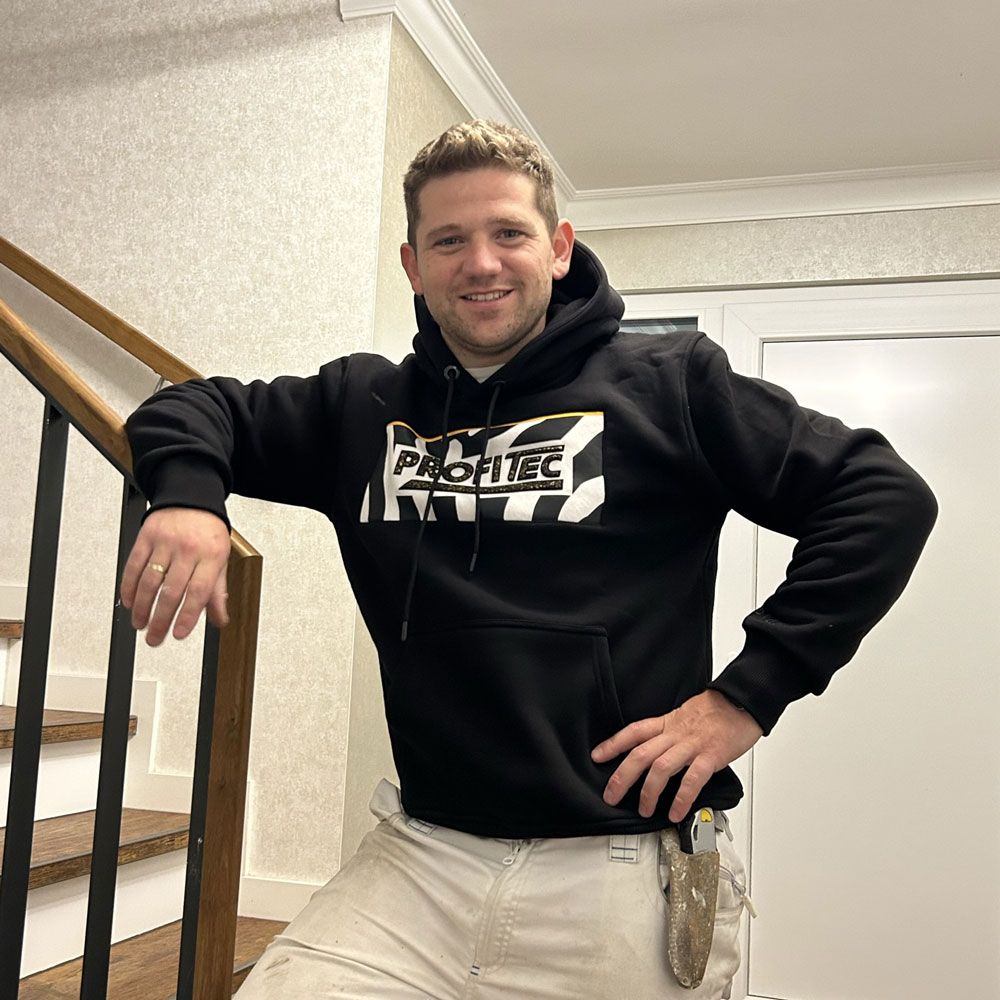 Ein Mann in schwarzem Kapuzenpulli und Khakihose lehnt mit einer Hand in der Hüfte an einer Holztreppe.