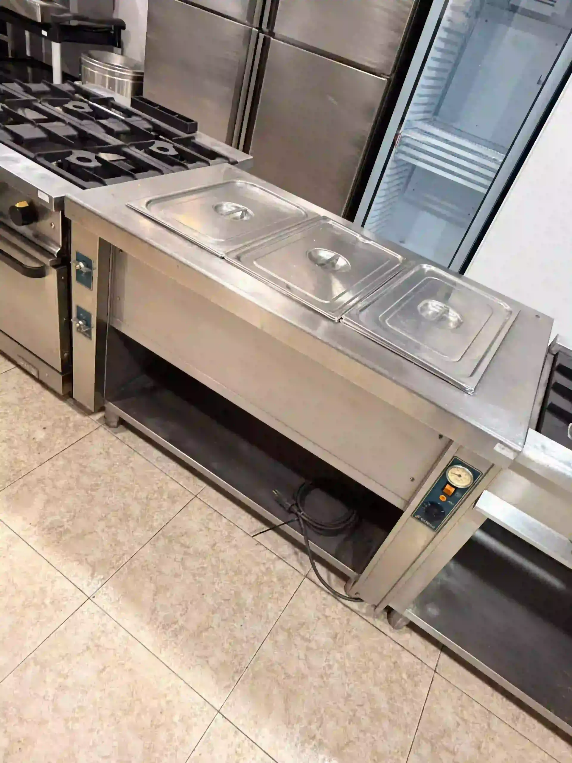 Encimera de cocina comercial de acero inoxidable con fregaderos y placas de cocina sobre suelo de baldosas.