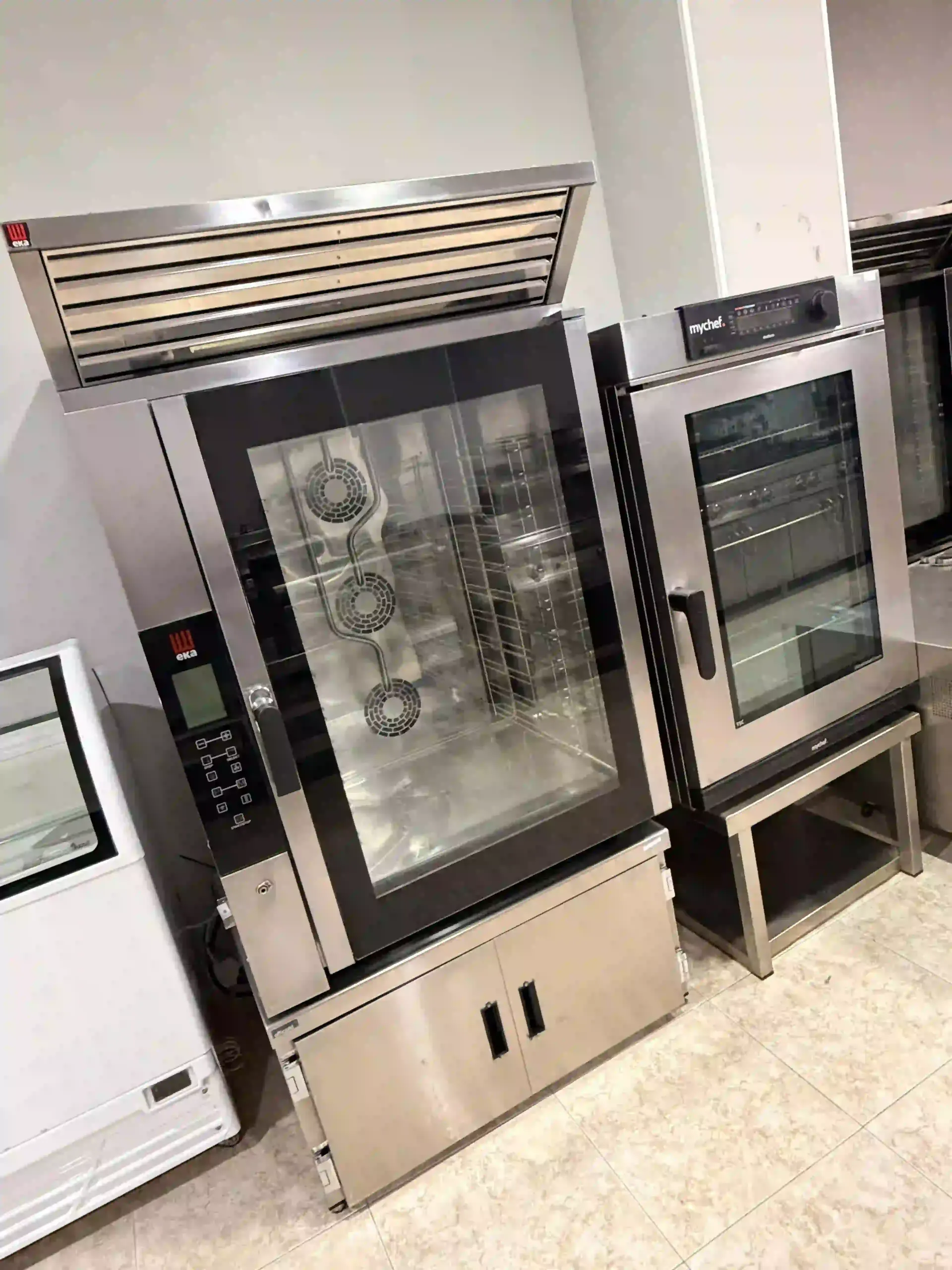 Hornos comerciales de acero inoxidable y armario calentador en una cocina, con puertas y estantes de vidrio.