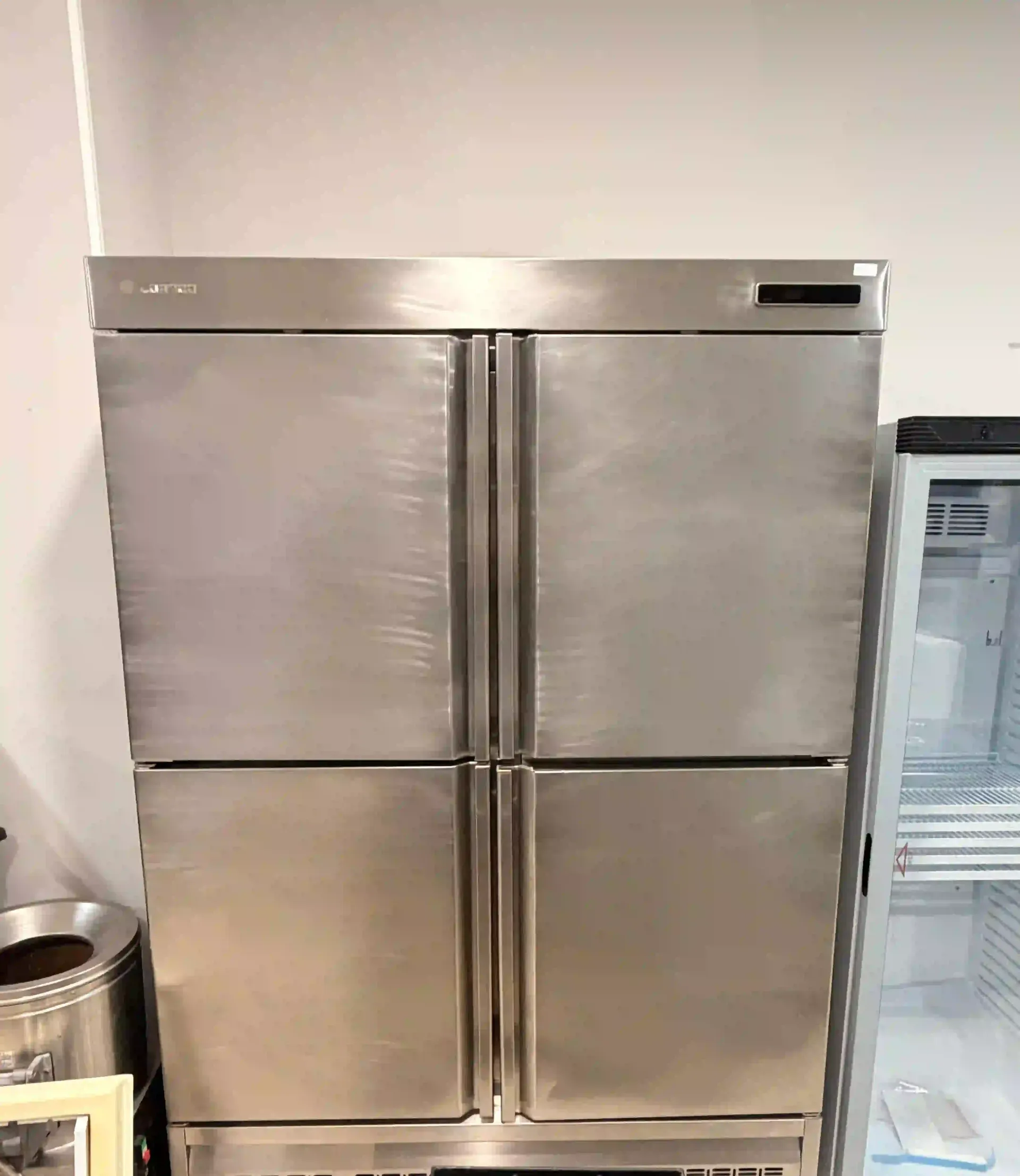 Frigorífico de cuatro puertas de acero inoxidable en una cocina o trastero, con un pequeño cubo de basura al lado.
