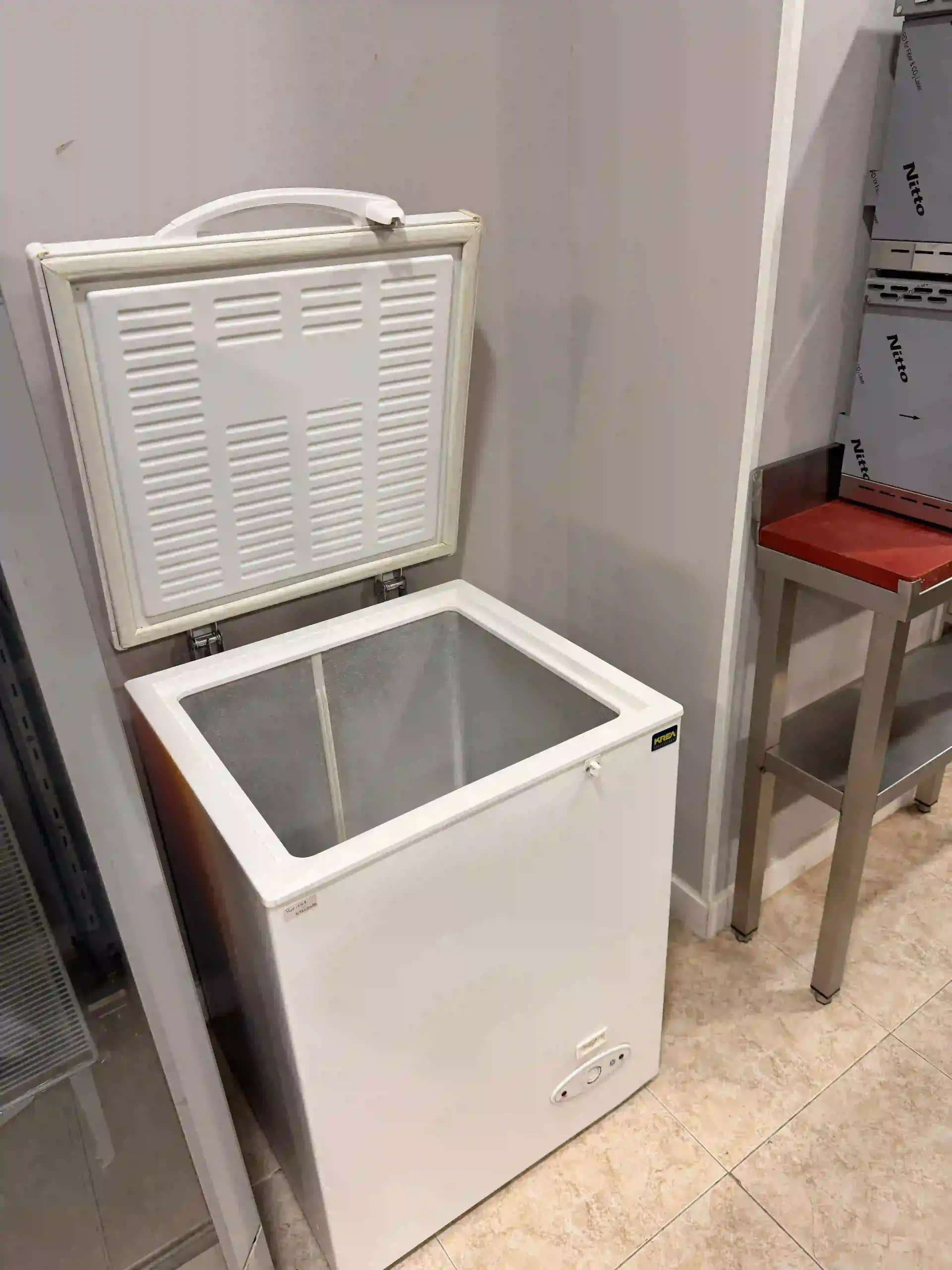 Congelador horizontal blanco con la tapa abierta, que deja ver su interior vacío en una habitación con suelo de baldosas.