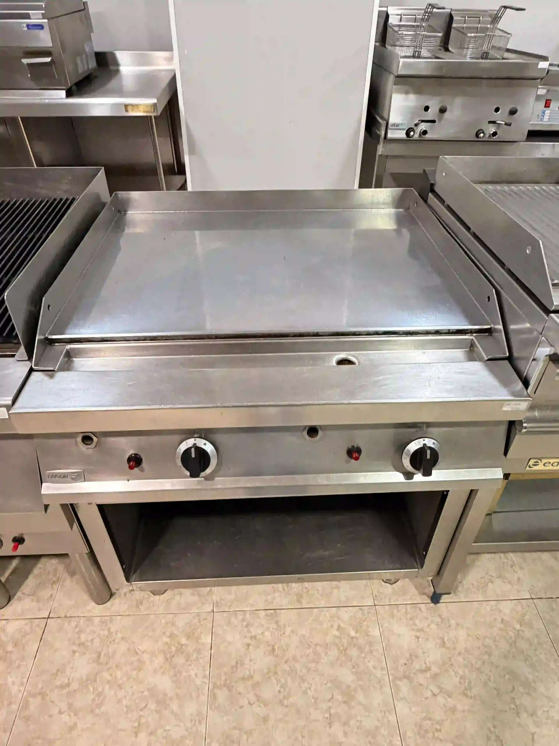 Plancha de acero inoxidable con superficie plana, equipada con mandos de control y estante inferior, en una cocina comercial.