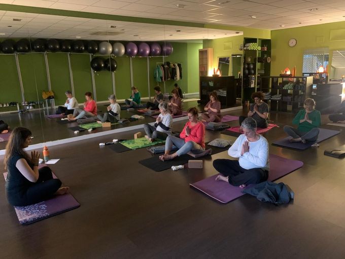 Clase de yoga: el instructor dirige posturas sentadas, estudiantes en colchonetas, estudio interior con espejos.