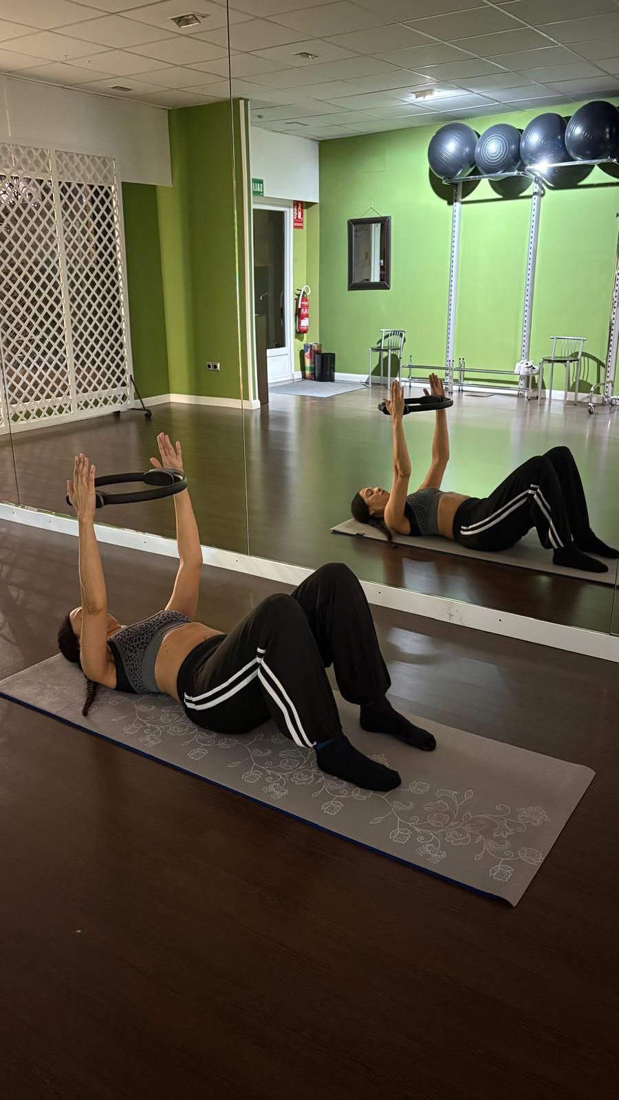 Mujer practicando pilates, tumbada sobre una colchoneta, sujetando una aro con los brazos en alto en el estudio.