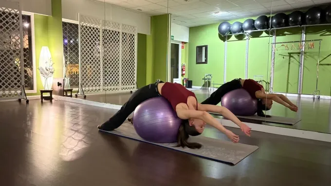 Mujer sobre una pelota de ejercicio morada, estirando la espalda en un estudio de fitness con espejos y paredes verdes.