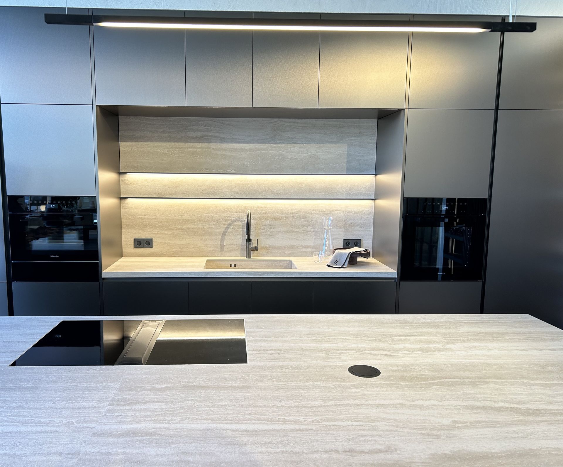 Cocina moderna con gabinetes grises, una isla central, electrodomésticos empotrados y un protector contra salpicaduras empotrado con iluminación.