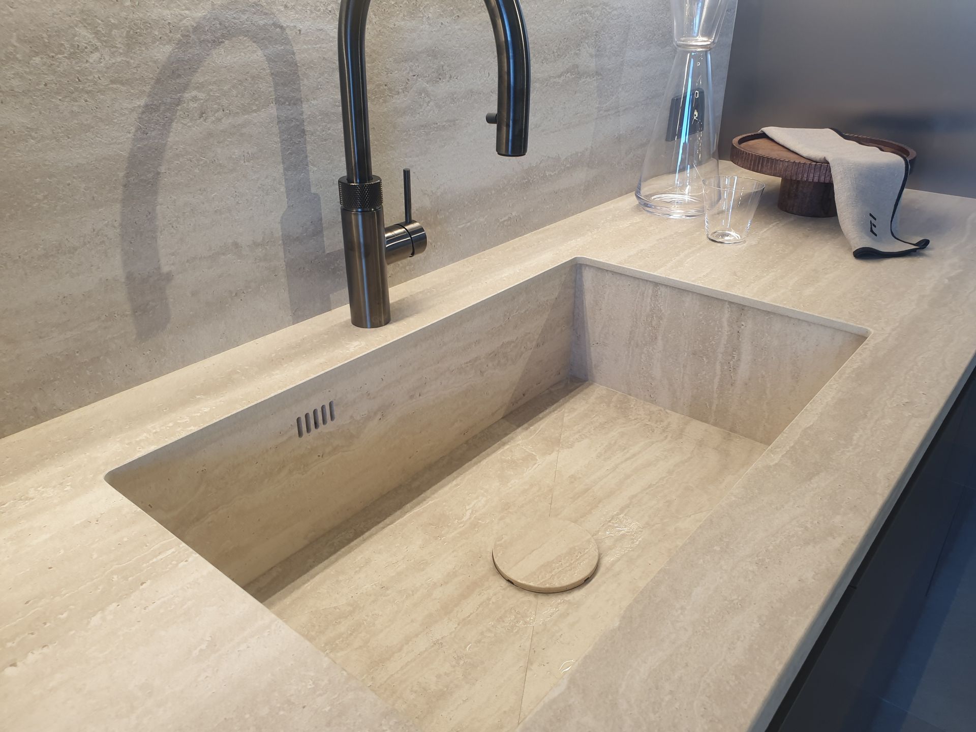 Un fregadero de cocina moderno hecho de piedra de color claro con un grifo negro, un desagüe y algunos elementos en la encimera.