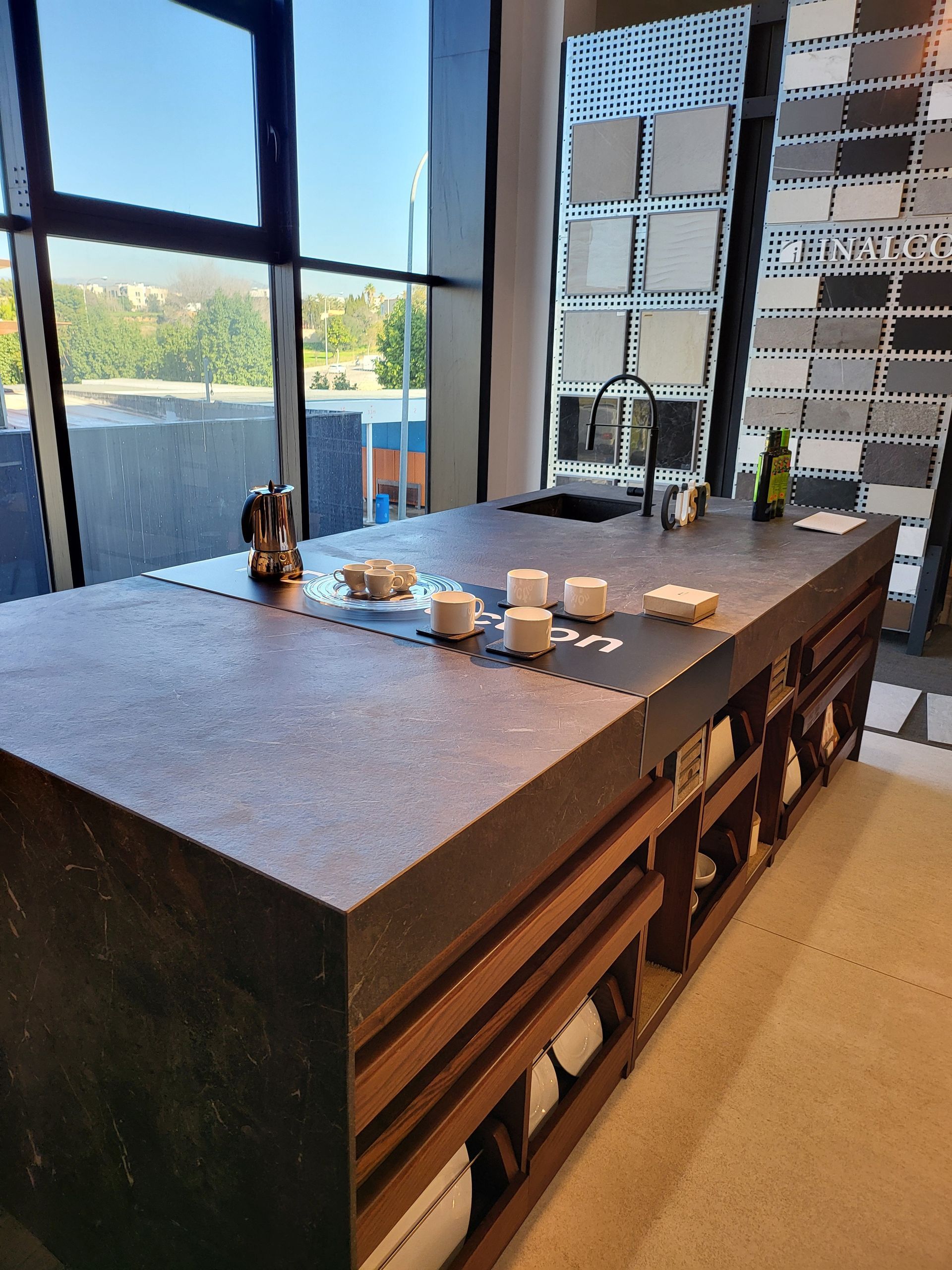 Isla de cocina con encimera oscura, estantes de madera y fregadero, con tazas y ventana al fondo.