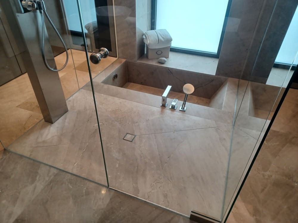 Baño con suelo de piedra, bañera incorporada, paredes de ducha de cristal y una ventana al fondo.