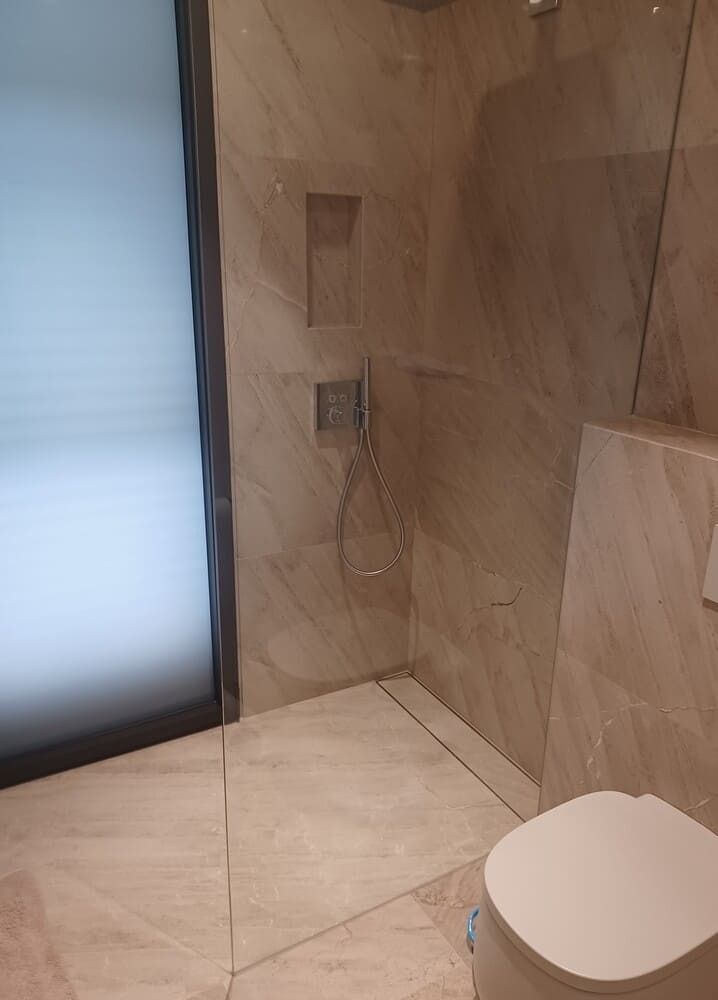 Baño con puerta de ducha de cristal, azulejos en tonos neutros, estante empotrado y un inodoro.