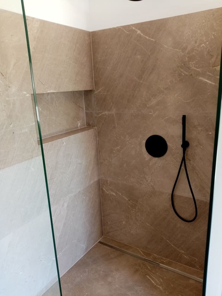 Ducha con azulejos de color beige, cabezal de ducha negro, puerta de cristal y estante incorporado.