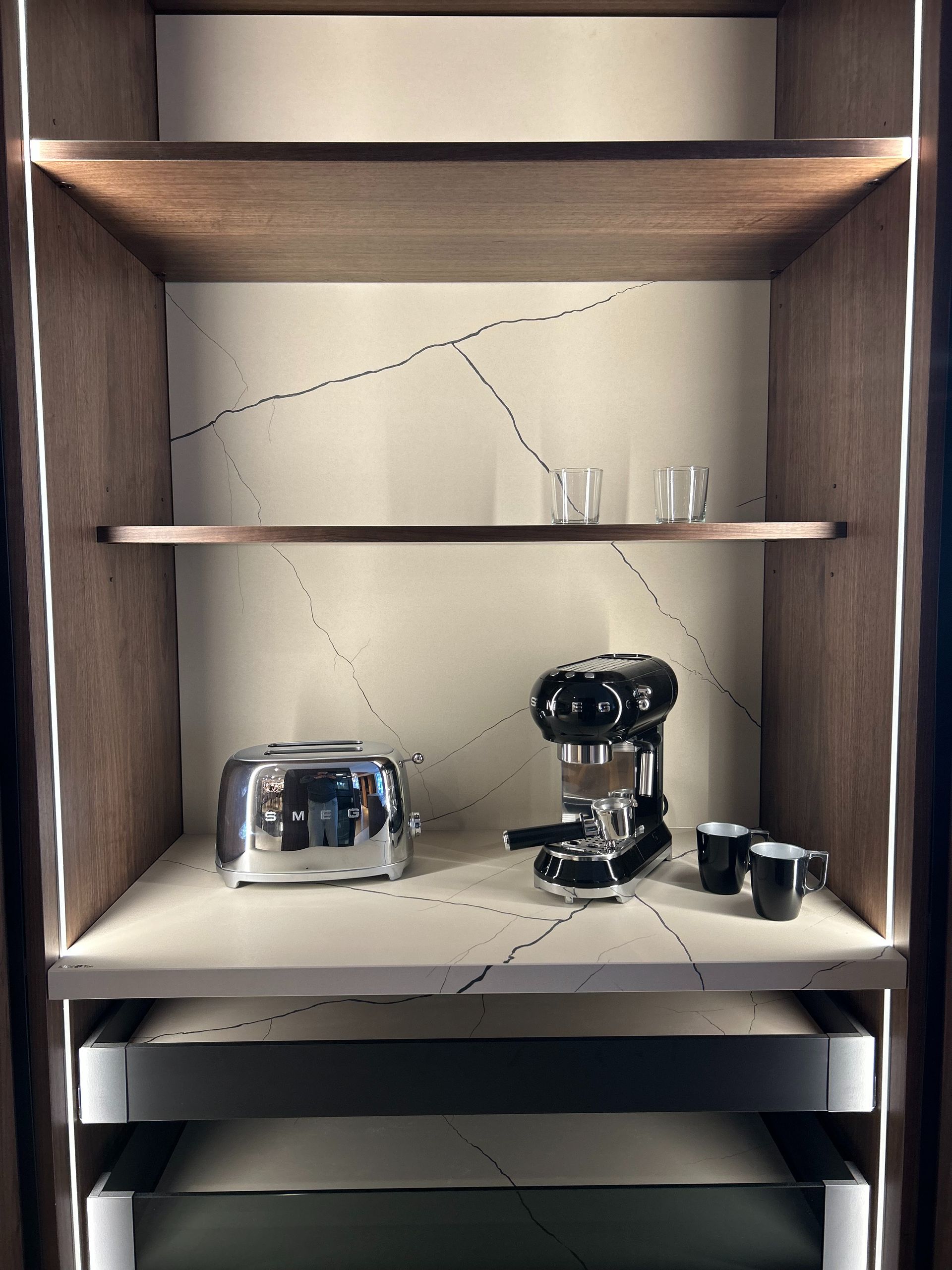 Un mueble con tostadora, cafetera expreso y tazas en estantes. El mueble tiene un diseño moderno e iluminación.