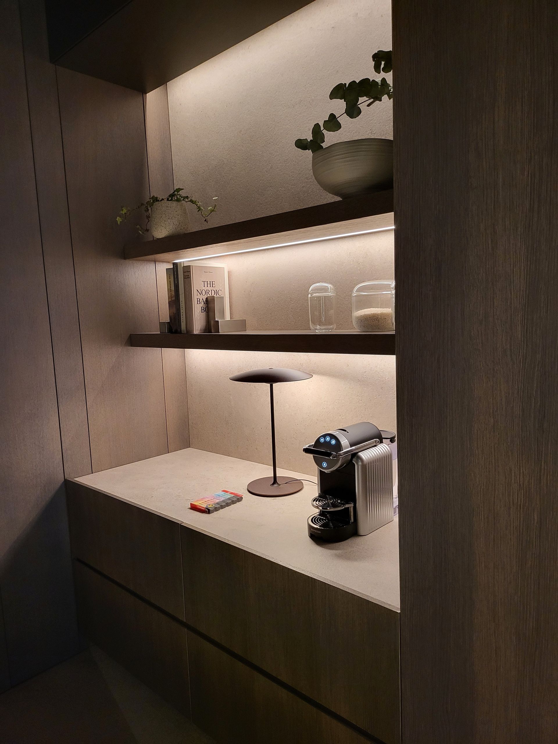 Estación de café con estantes, armarios empotrados e iluminación ambiental. Incluye cafetera y plantas pequeñas.