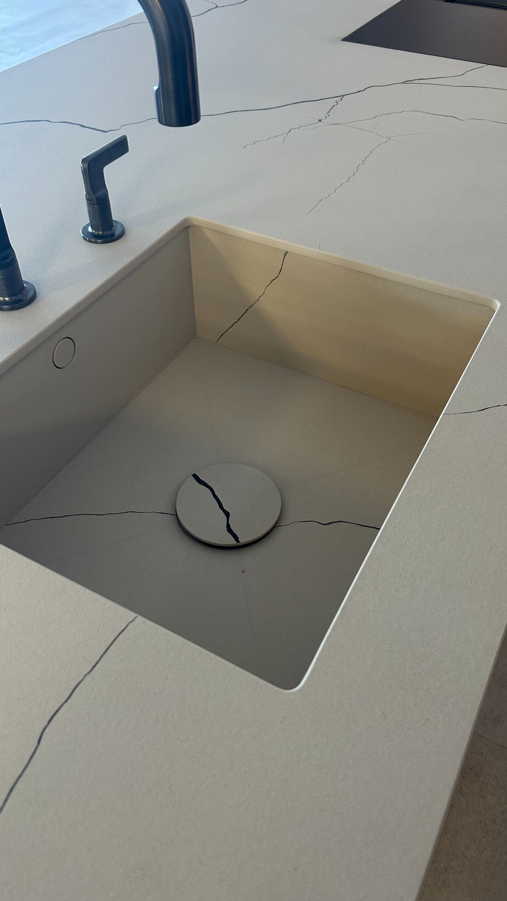 Un lavabo cuadrado beige sobre una encimera clara, con grifería negra. La tapa del desagüe está rota.