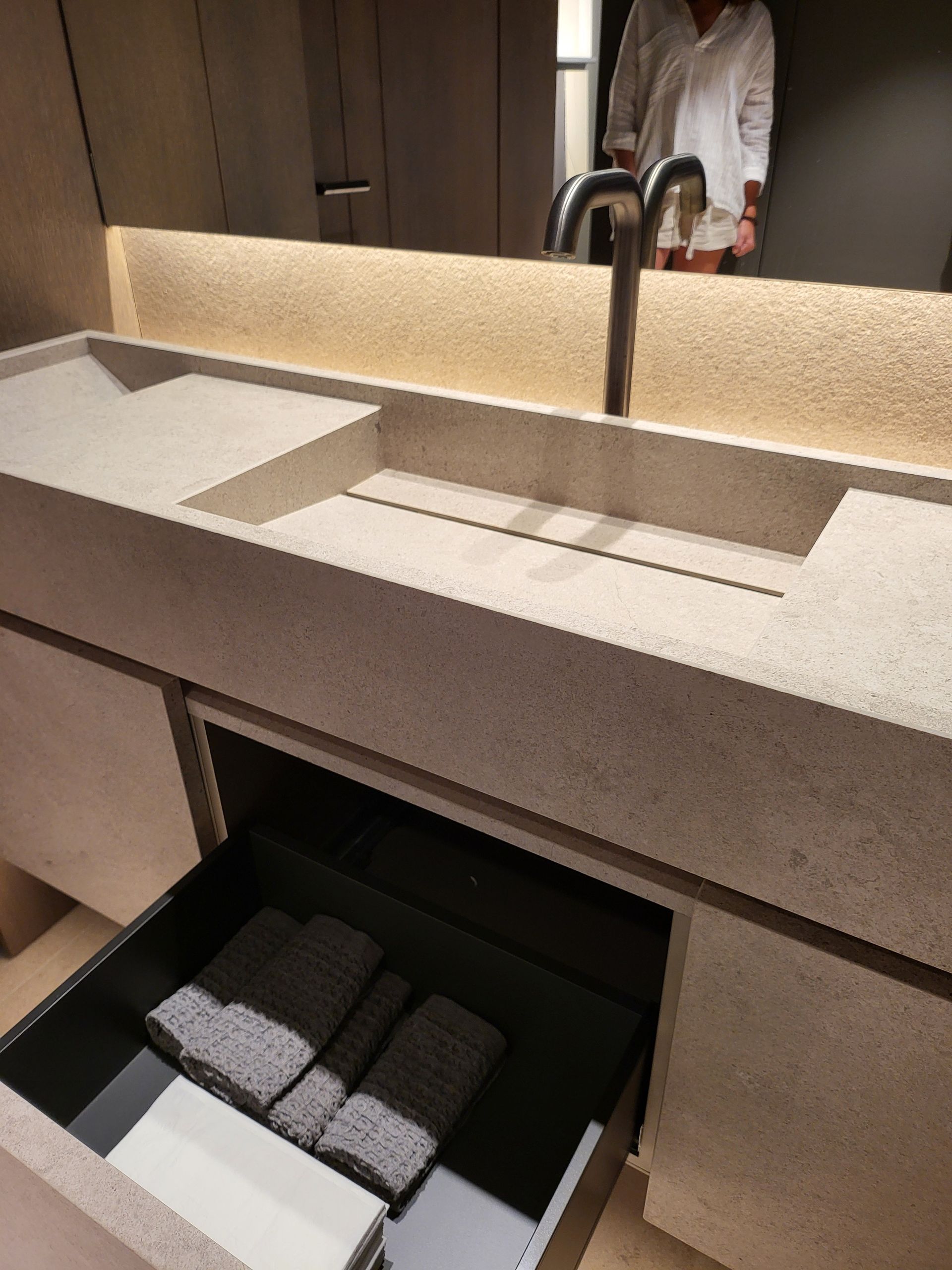 Mueble de baño con cajón abierto. Lavabo de piedra, toallas y una persona reflejada en el espejo.