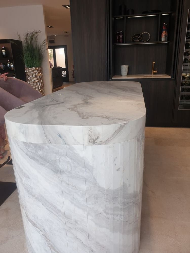 Isla de mármol en la cocina. Muebles oscuros, grifo dorado y estantes con botellas.