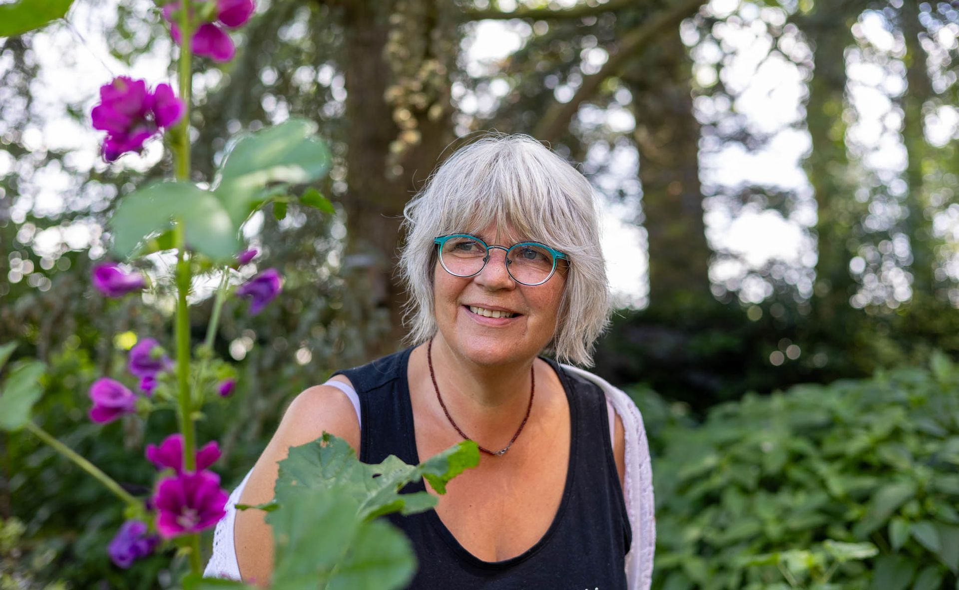 Eine Frau mit Brille steht vor lila Blumen in einem Garten.