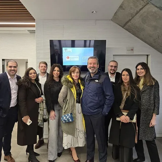 Grupo de personas posando cerca de una pantalla en un edificio moderno. Algunas llevan abrigos.
