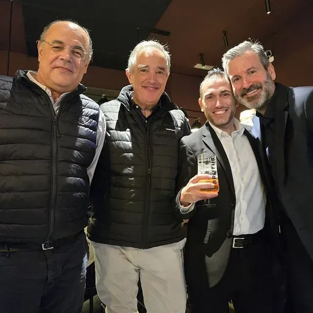 Cuatro hombres sonrientes, en el interior. Uno sostiene un vaso de cerveza. Dos llevan chaquetas negras, los otros, camisas. Luz tenue.