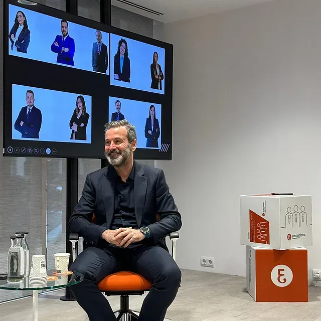Un hombre con traje está sentado; una pantalla muestra a otros. En la habitación hay cajas y una mesa de cristal.