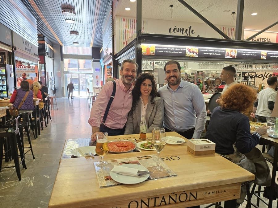 Tres personas sonriendo en una mesa en un mercado de alimentos, con comida y bebidas a la vista.