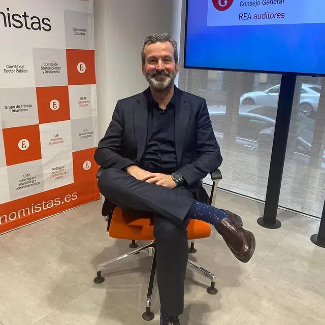 Un hombre con traje está sentado con las piernas cruzadas. Está delante de un cartel que dice 