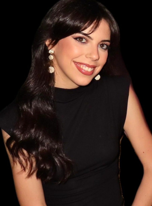 Una persona sonriente con cabello largo, oscuro y ondulado, que lleva una blusa negra sin mangas y pendientes blancos colgantes sobre un fondo negro.