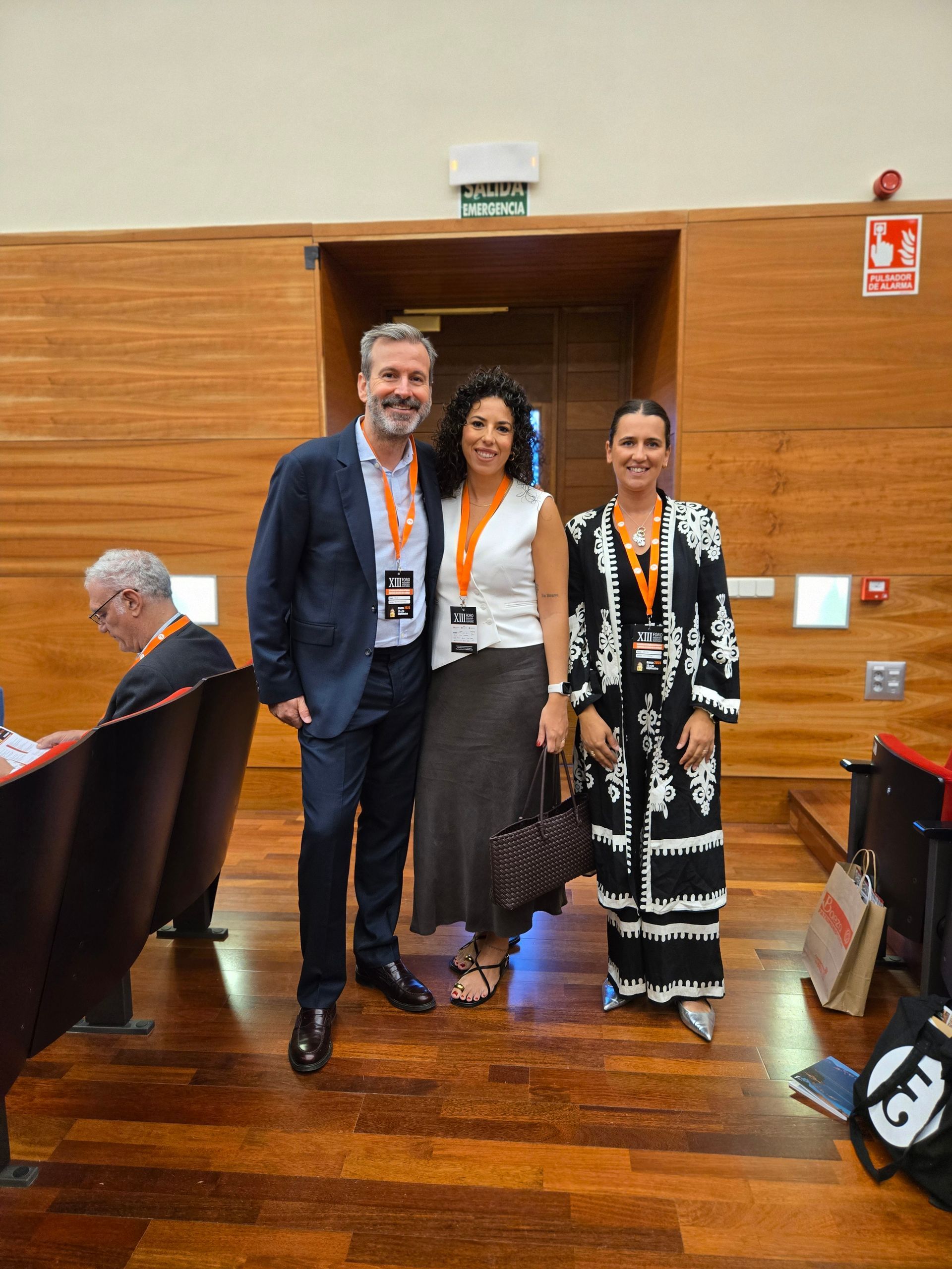 Tres personas con acreditaciones están de pie dentro de un edificio. Un hombre con traje y dos mujeres, una con blusa y falda blancas, la otra con un vestido negro estampado.