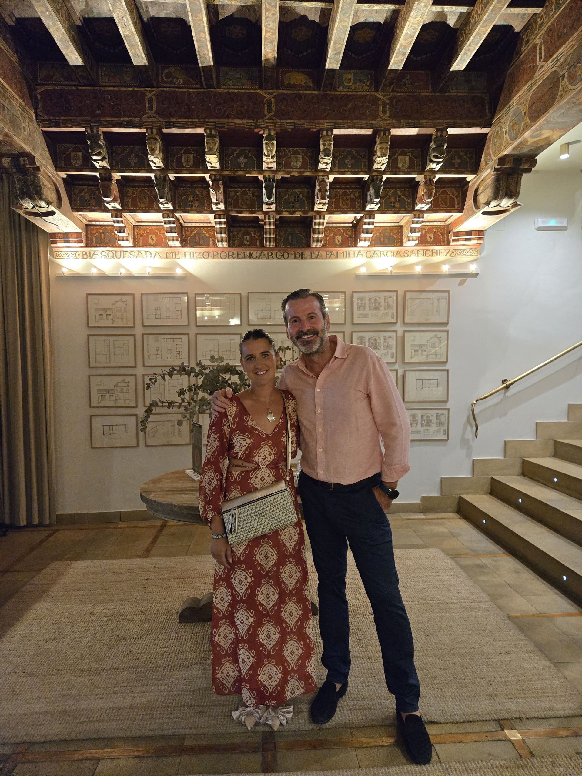 Dos personas posando en un edificio con techo decorado, bocetos en la pared y escaleras. Una de ellas lleva un vestido de flores.
