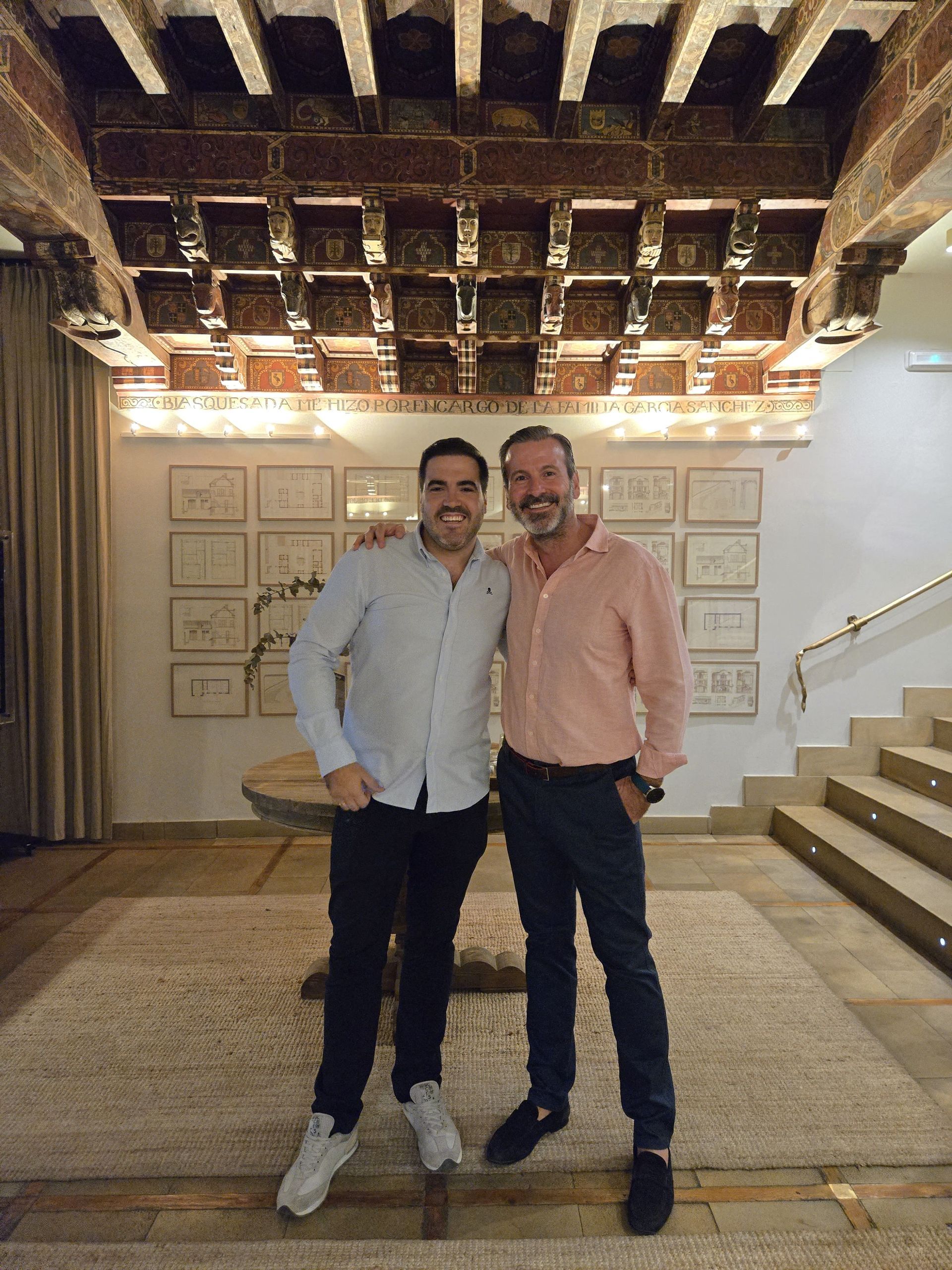 Dos hombres sonrientes, posando juntos dentro de un edificio con un techo decorado y dibujos en la pared.