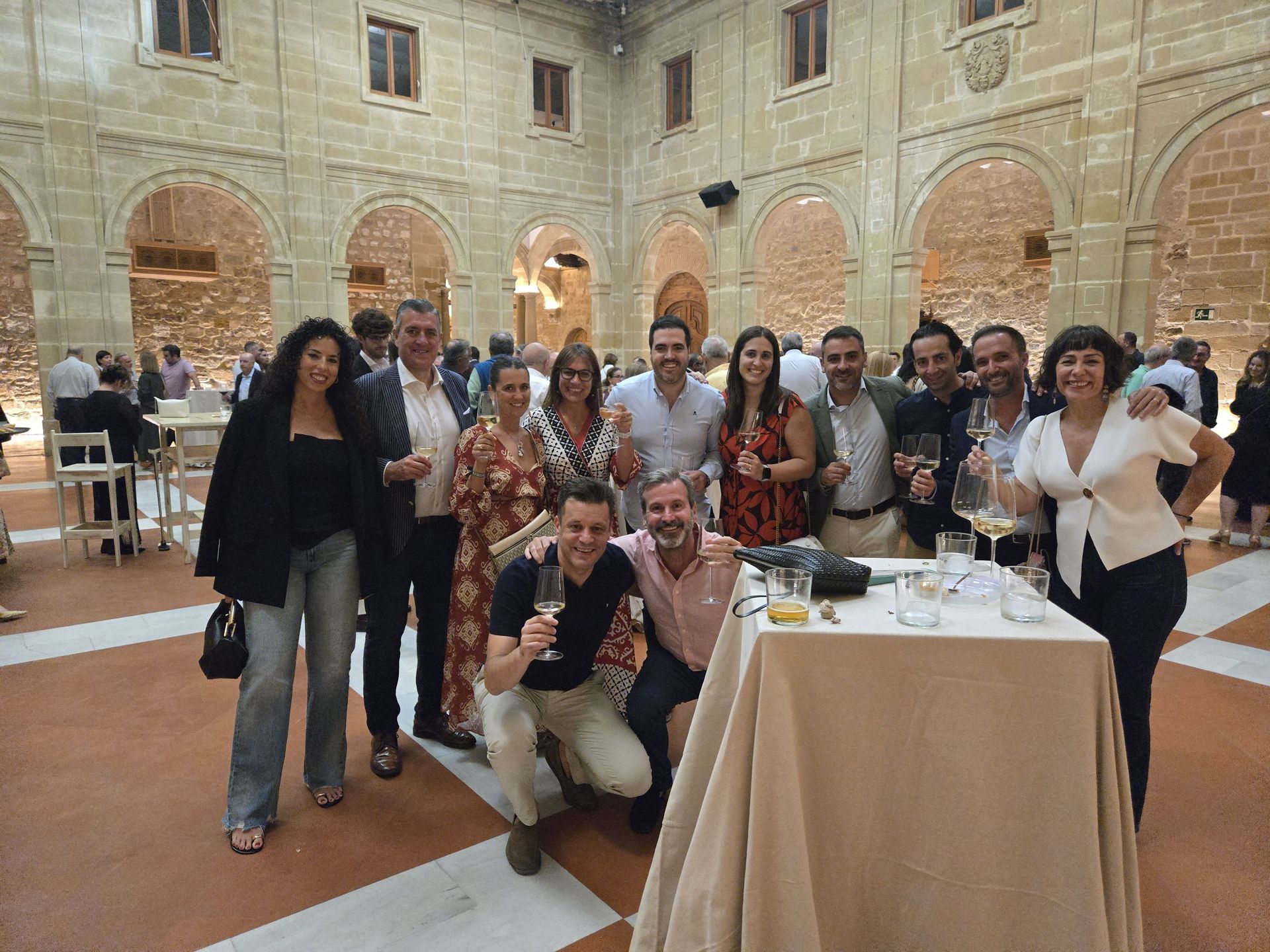 Grupo de personas celebrando, algunas con copas en mano, en un patio con arcos de piedra.
