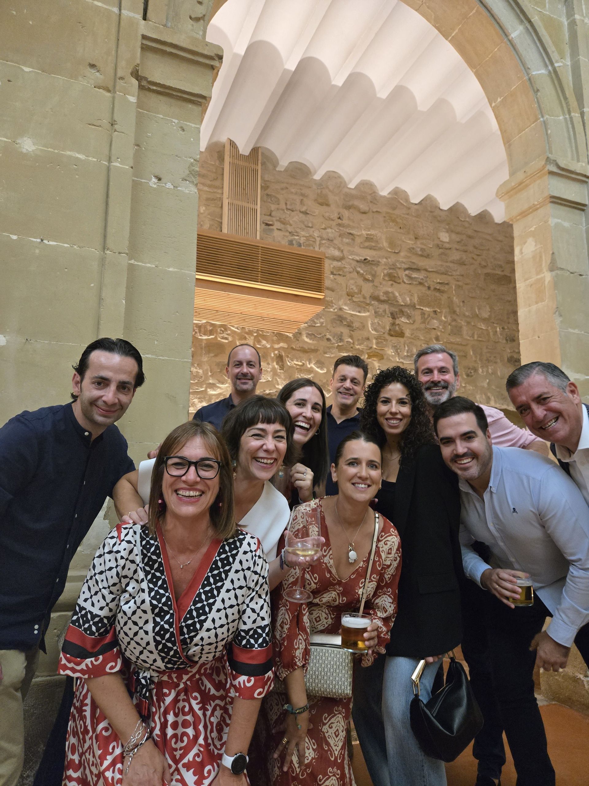 Grupo de personas sonrientes, posando frente a un arco de piedra. Algunos llevan bebidas.