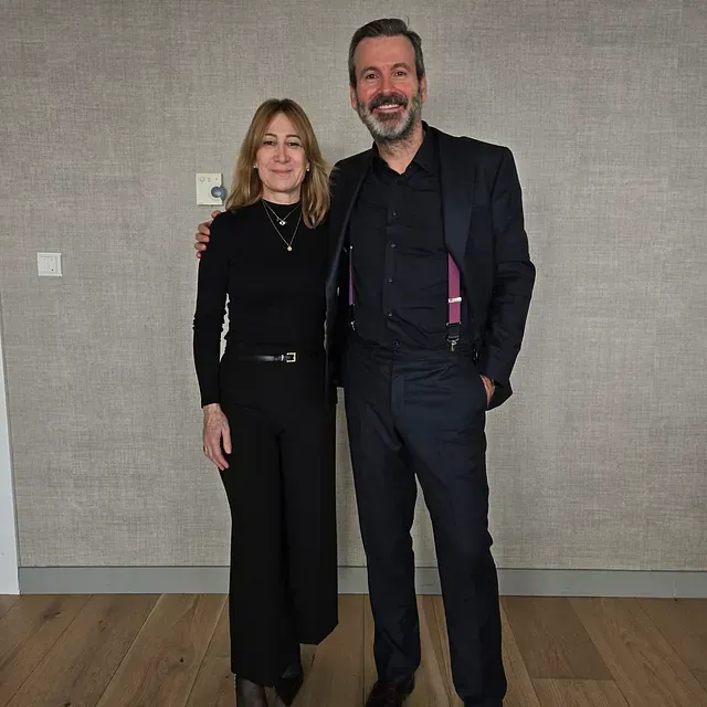 Una mujer con traje pantalón negro y un hombre con traje posan juntos, sonriendo, en el interior.