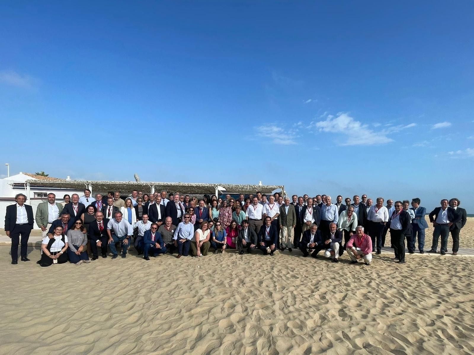 Un numeroso grupo de personas en una playa frente a un edificio blanco, bajo un cielo azul despejado.