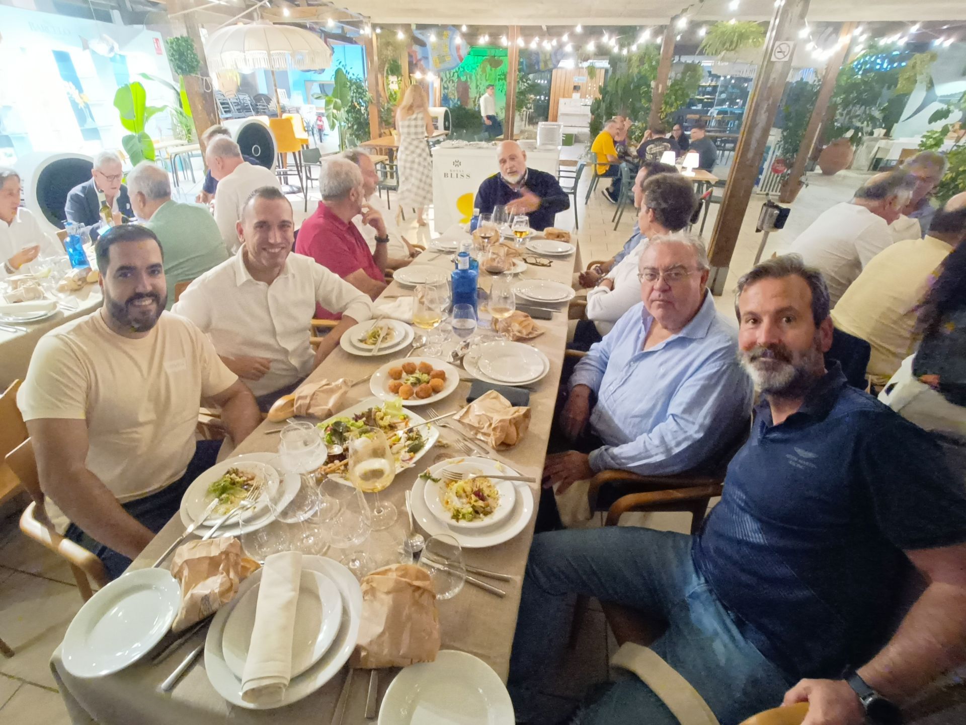 Gente comiendo y charlando en una mesa al aire libre de un restaurante. Platos de comida, pan y bebidas. Guirnaldas de luces en el techo.