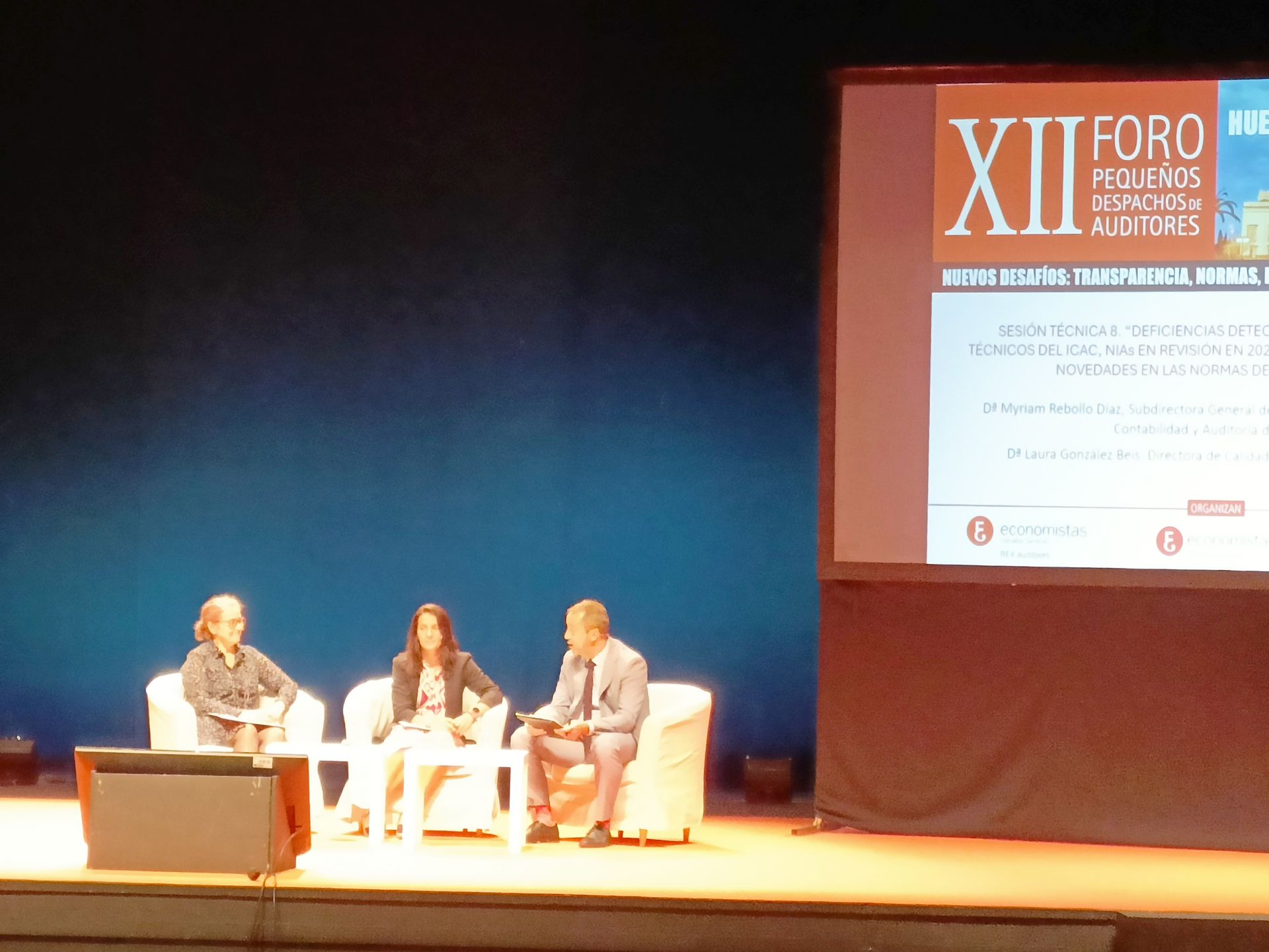 Mesa redonda en el escenario con tres personas, pantalla de fondo y público.