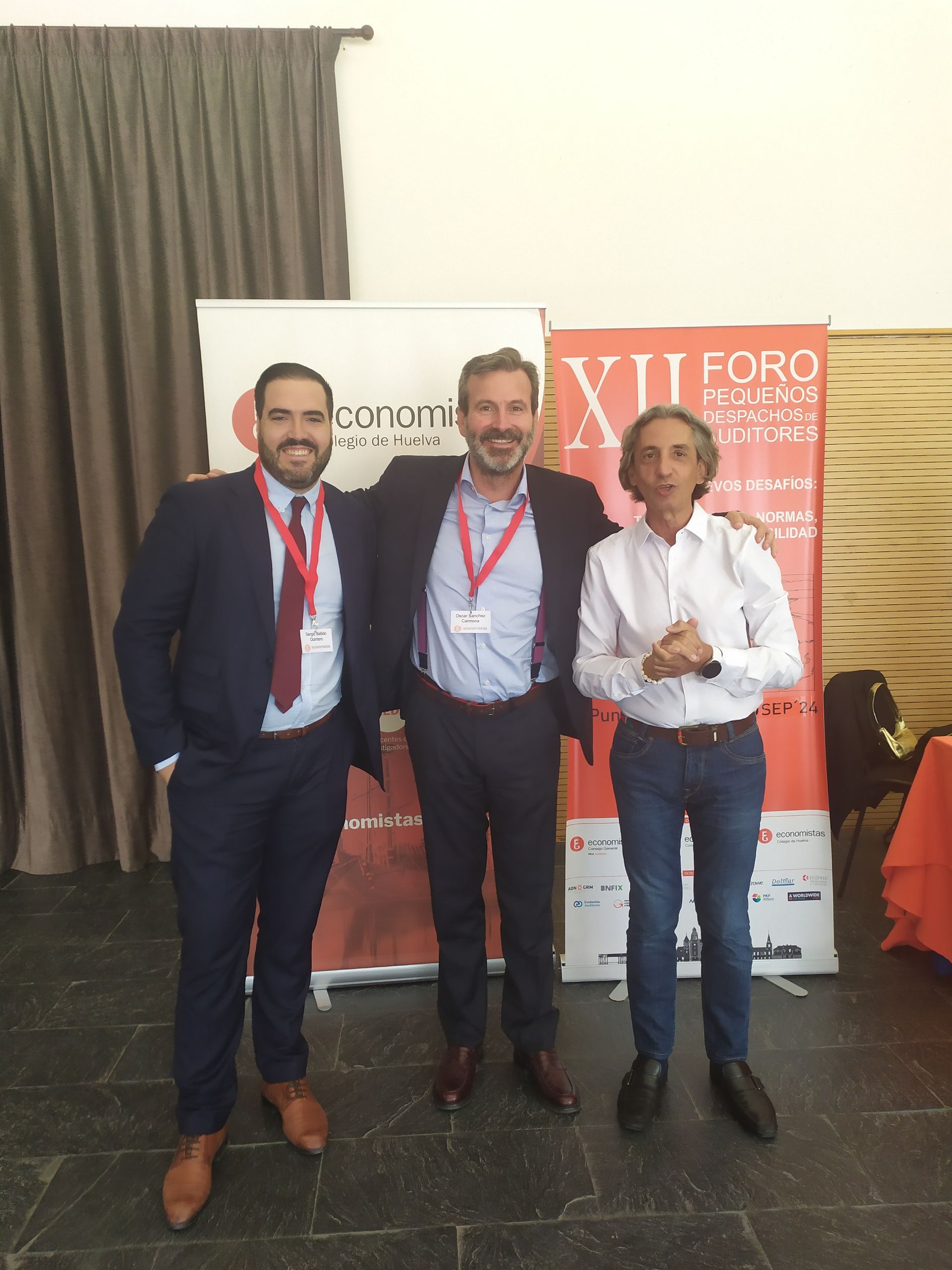 Tres hombres están juntos frente a una pancarta. Visten trajes y sonríen, posando para una foto.