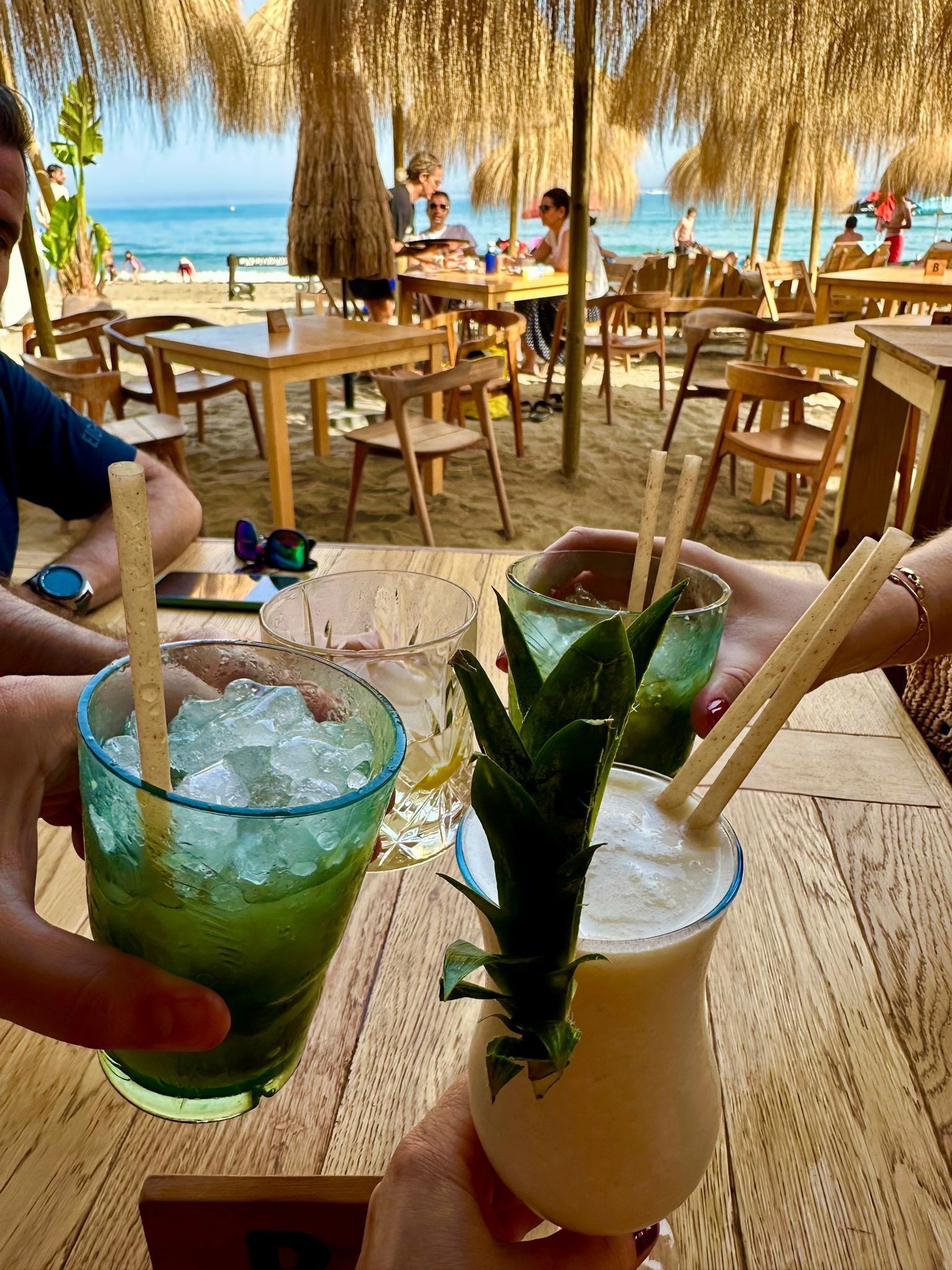 Gente brindando con cócteles en un restaurante junto a la playa, sombrilla de paja, mesas y el océano al fondo.