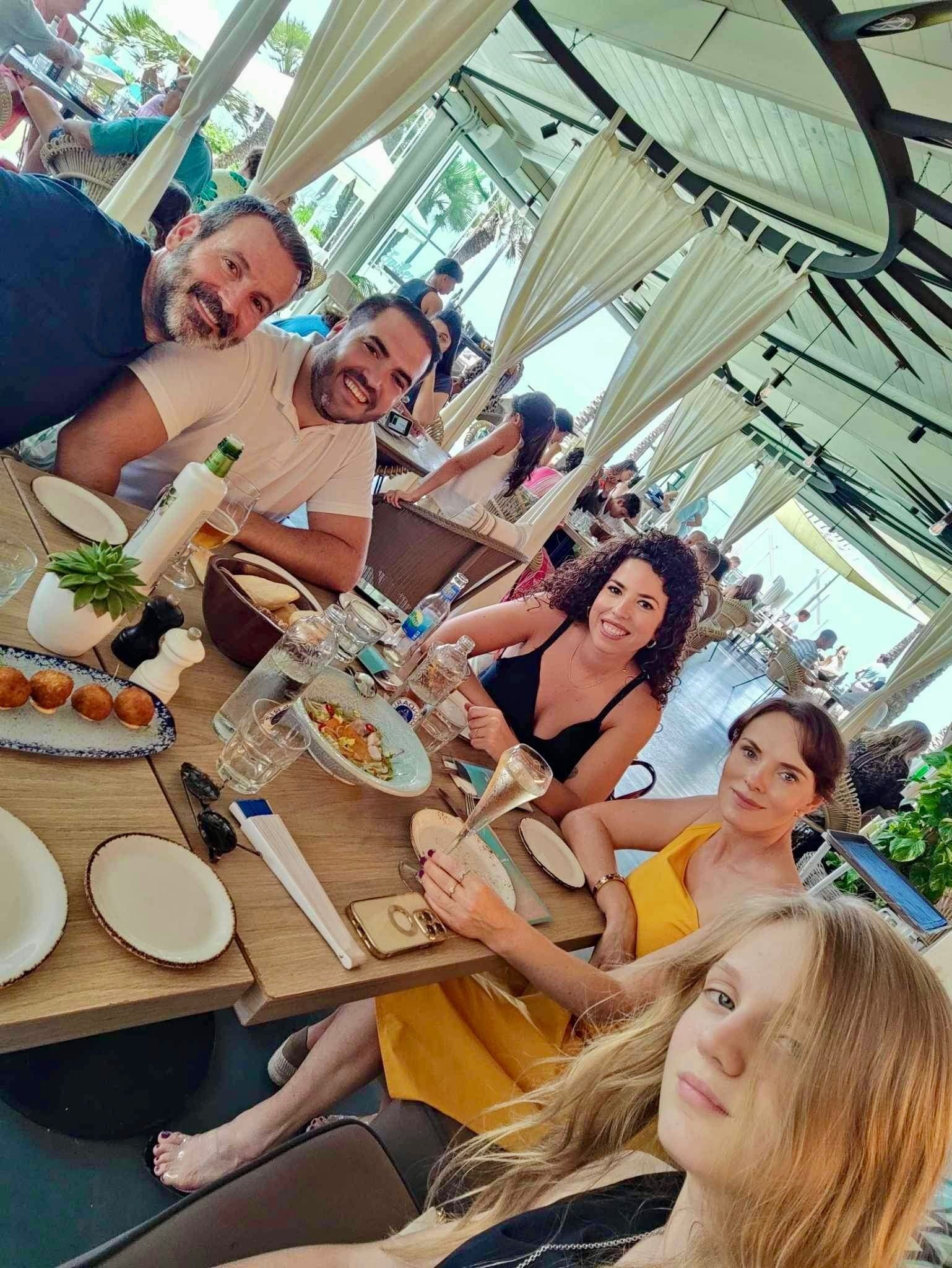 Personas sentadas a la mesa de un restaurante, platos de comida. Bajo un toldo blanco.