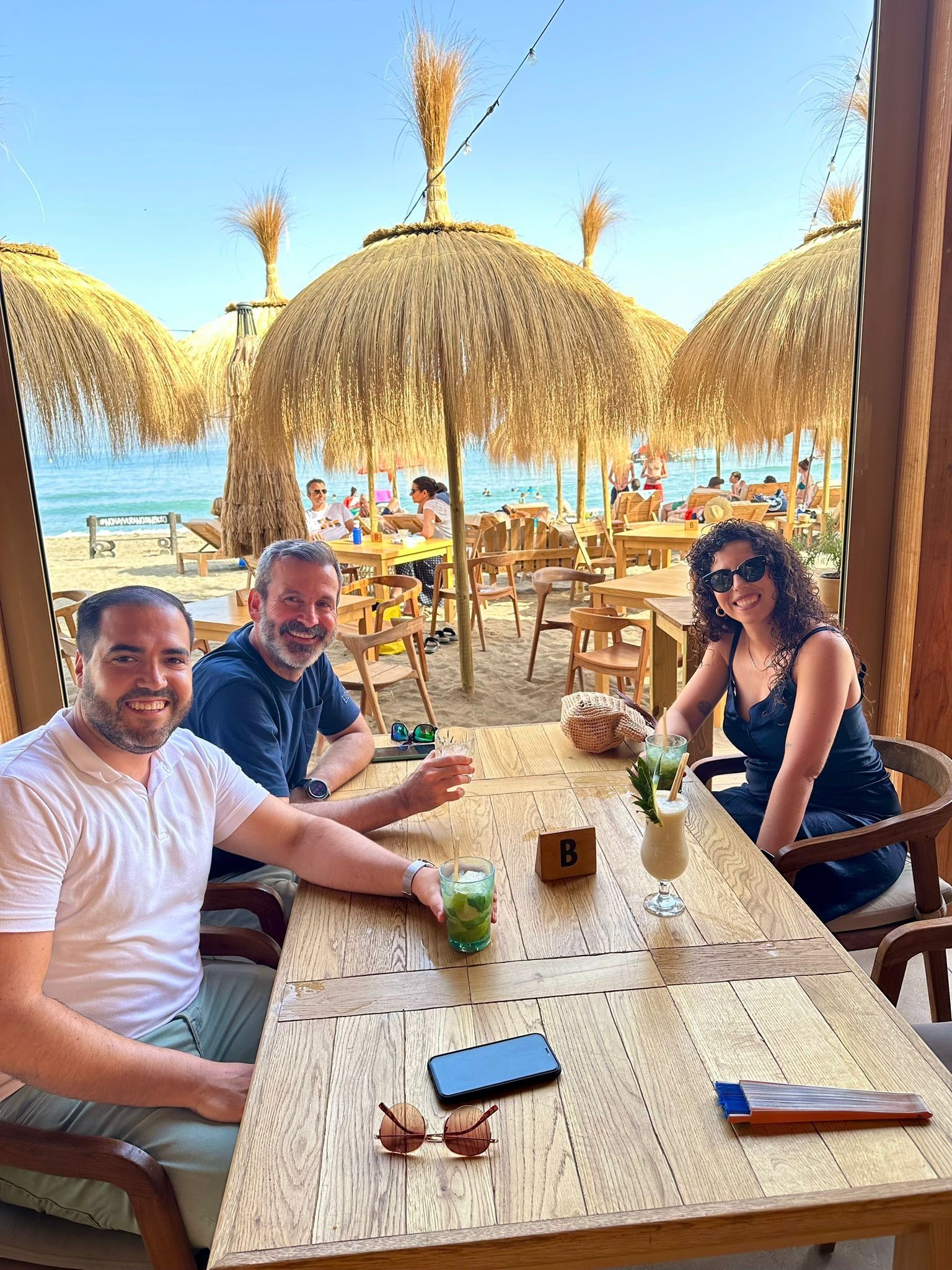 Tres personas sentadas a una mesa en la playa con bebidas. Sombrillas de playa, fondo oceánico.