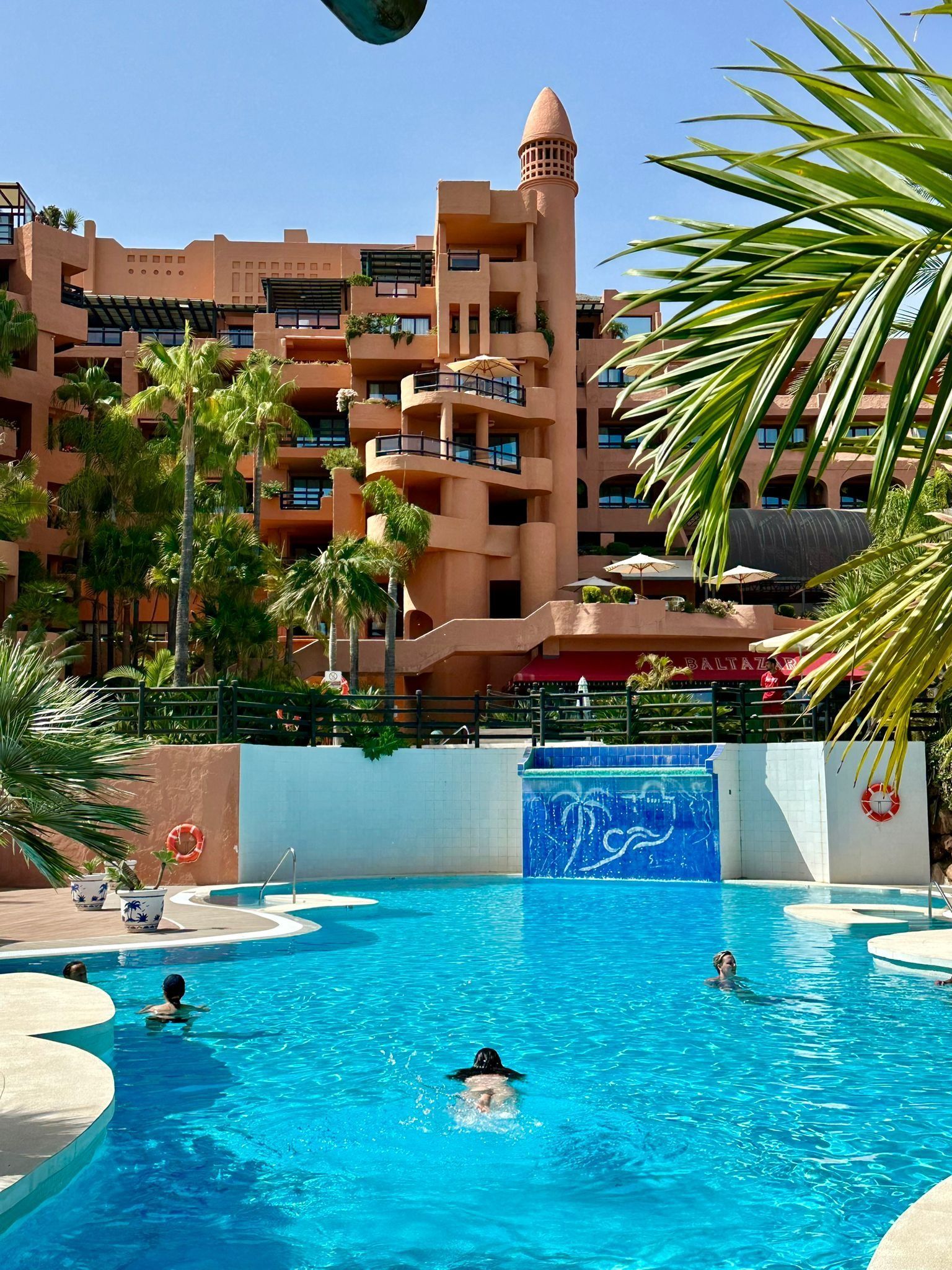 Piscina frente a un edificio de terracota de varias plantas. Gente nadando en el agua azul. Palmeras. Día soleado.