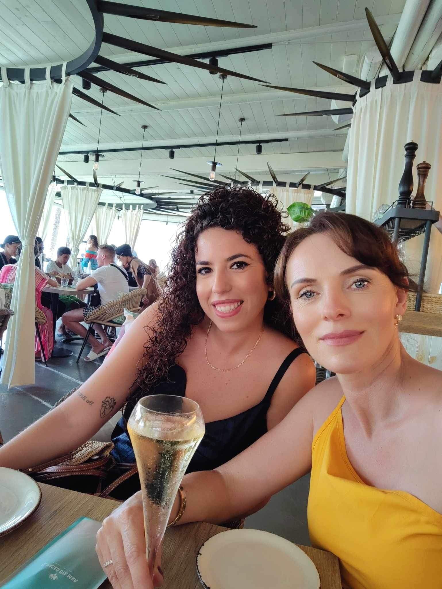 Dos mujeres sonriendo a la cámara, una de ellas sosteniendo una botella de champán, sentadas en un restaurante con otras personas.