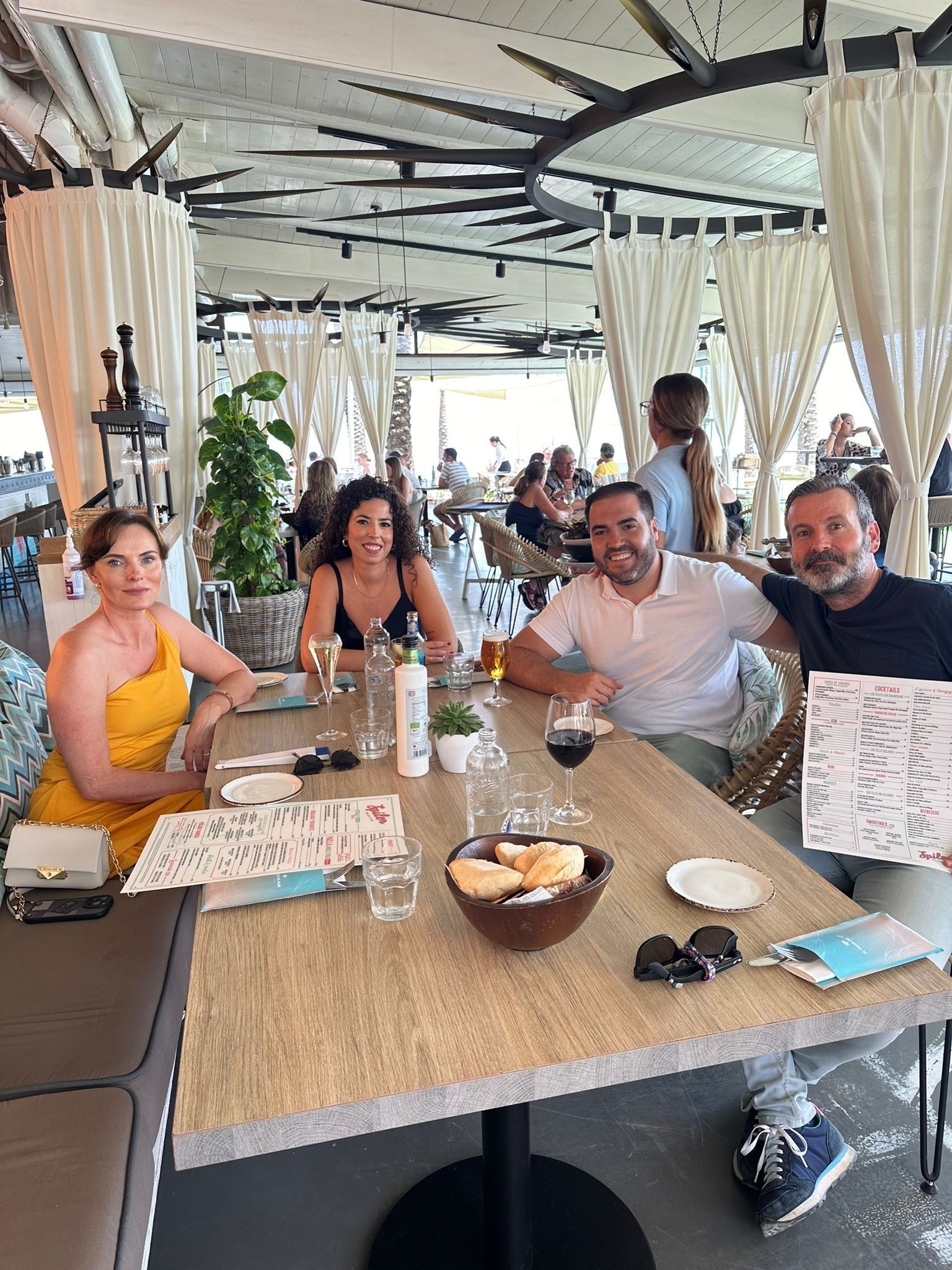 Personas sonrientes sentadas a la mesa de un restaurante. Mujer con vestido amarillo. Mesa de madera con comida y bebidas. Ambiente al aire libre.