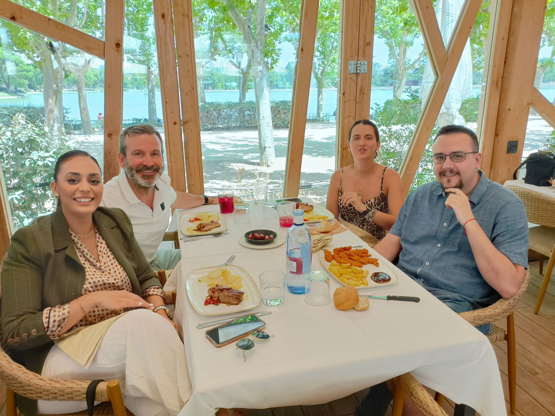 Cuatro personas sentadas a una mesa, comiendo. Dentro de una estructura con armazón de madera, con vistas al agua.